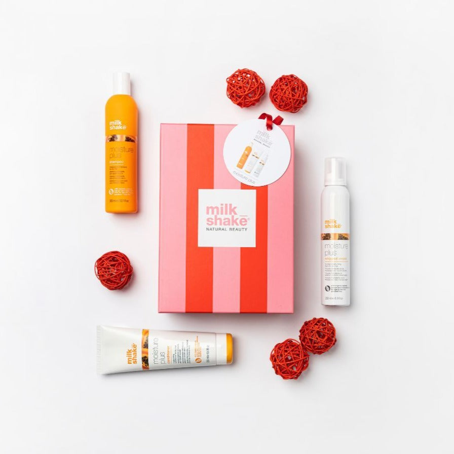 Milk Shake Gift Set with Moisture Plus Trio, including a shampoo, conditioner, and hair treatment spray, arranged alongside a red and pink striped gift box and decorative red balls on a white background.